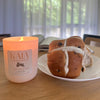Hot cross bun with white icing on a plate next to a lit candle labeled 'Kama' on a wooden surface.