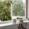 Candle with 'KAIA' branding on a windowsill with a view of greenery outside.