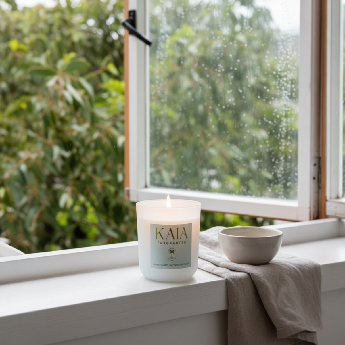 Candle with 'KAIA' branding on a windowsill with a view of greenery outside.
