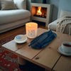 Candle labeled 'KAIA' with candle care kit on a wooden table with a cozy living room background